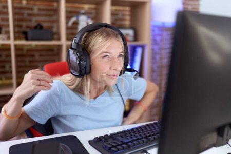 Téléchargez les photos : Jeune femme caucasienne jouant à des jeux vidéo portant des écouteurs souffrant de maux de dos, toucher le dos avec la main, douleurs musculaires - en image libre de droit
