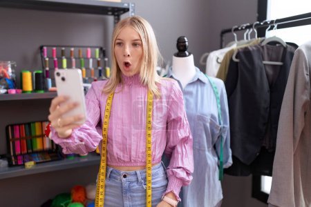 Foto de Young caucasian woman dressmaker designer on video call with smartphone scared and amazed with open mouth for surprise, disbelief face - Imagen libre de derechos
