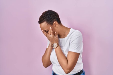 Schöne afrikanisch-amerikanische Frau steht vor rosa Hintergrund mit traurigem Gesichtsausdruck, der das Gesicht mit Händen bedeckt, während sie weint. Depressionskonzept. 