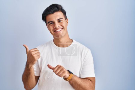 Photo for Hispanic man standing over blue background pointing to the back behind with hand and thumbs up, smiling confident - Royalty Free Image