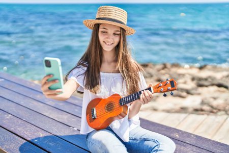 Foto de Adorable girl tourist make selfie by the smartphone playing ukulele at seaside - Imagen libre de derechos