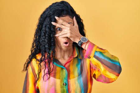 Foto de Mujer hispana de mediana edad de pie sobre fondo amarillo asomándose en shock cubriendo la cara y los ojos con la mano, mirando a través de los dedos con expresión avergonzada. - Imagen libre de derechos