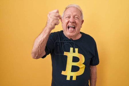 Foto de Senior man with grey hair wearing bitcoin t shirt angry and mad raising fist frustrated and furious while shouting with anger. rage and aggressive concept. - Imagen libre de derechos