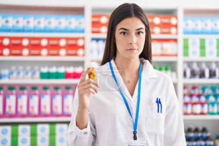 Photo for Hispanic woman working at pharmacy drugstore holding pills thinking attitude and sober expression looking self confident - Royalty Free Image
