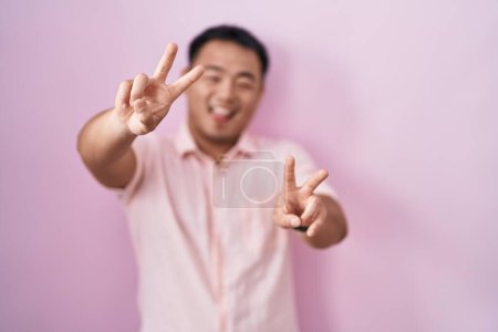 Foto de Joven chino de pie sobre fondo rosa sonriendo con la lengua hacia fuera mostrando los dedos de ambas manos haciendo signo de victoria. número dos. - Imagen libre de derechos