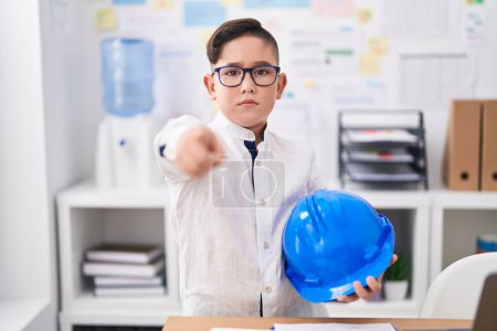 Photo for Young hispanic kid holding architect hardhat at the office pointing with finger to the camera and to you, confident gesture looking serious - Royalty Free Image