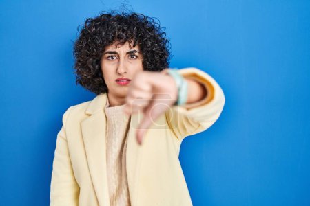 Foto de Young brunette woman with curly hair standing over blue background looking unhappy and angry showing rejection and negative with thumbs down gesture. bad expression. - Imagen libre de derechos