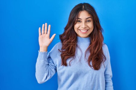 Téléchargez les photos : Jeune femme hispanique debout sur fond bleu renonçant à dire bonjour heureux et souriant, geste de bienvenue amical - en image libre de droit