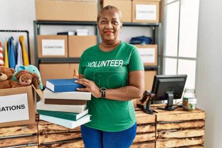 Photo for Senior african american woman wearing volunteer uniform holding books at charity center - Royalty Free Image
