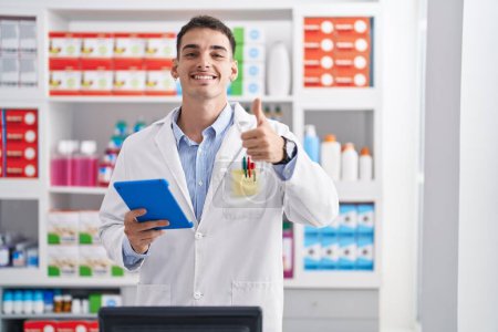 Photo for Handsome hispanic man working at pharmacy drugstore with tablet smiling happy and positive, thumb up doing excellent and approval sign - Royalty Free Image