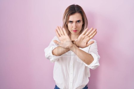 Téléchargez les photos : Young beautiful woman standing over pink background rejection expression crossing arms and palms doing negative sign, angry face - en image libre de droit