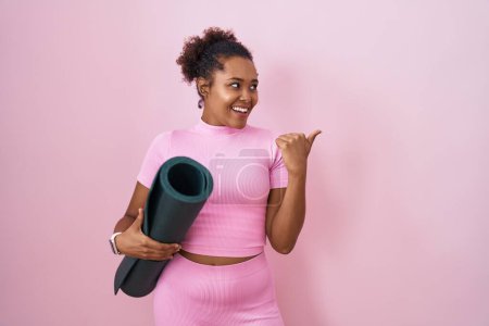 Téléchargez les photos : Jeune femme hispanique avec les cheveux bouclés tenant tapis de yoga sur fond rose souriant avec le visage heureux regardant et pointant vers le côté avec le pouce vers le haut. - en image libre de droit