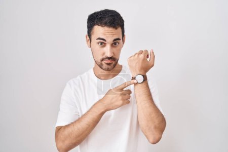Téléchargez les photos : Handsome hispanic man standing over white background in hurry pointing to watch time, impatience, looking at the camera with relaxed expression - en image libre de droit