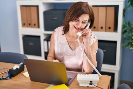 Foto de Mujer de mediana edad trabajadora de negocios hablando por teléfono usando el ordenador portátil en la oficina - Imagen libre de derechos