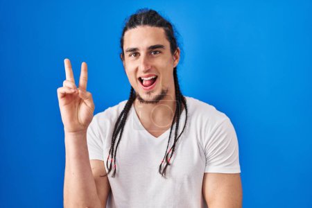 Photo for Hispanic man with long hair standing over blue background smiling with happy face winking at the camera doing victory sign. number two. - Royalty Free Image