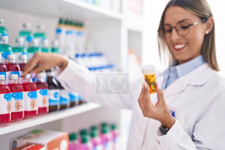 Foto de Joven hermosa mujer hispana farmacéutico sonriendo confiado sosteniendo pastillas botella en la farmacia - Imagen libre de derechos