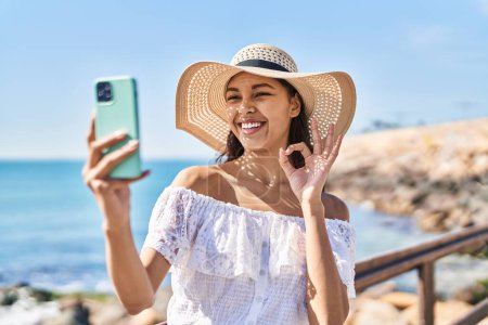 Foto de Mujer brasileña joven tomando una foto selfie con teléfono inteligente al aire libre haciendo signo de ok con los dedos, sonriendo gesto amistoso excelente símbolo - Imagen libre de derechos