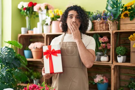 Foto de Hombre hispano con el pelo rizado trabajando en floristería sosteniendo regalo cubriendo la boca con la mano, sorprendido y temeroso de equivocarse. expresión sorprendida - Imagen libre de derechos