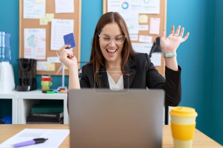 Téléchargez les photos : Jeune belle travailleuse d'affaires utilisant un ordinateur portable tenant une carte de crédit avec expression de célébration au bureau - en image libre de droit