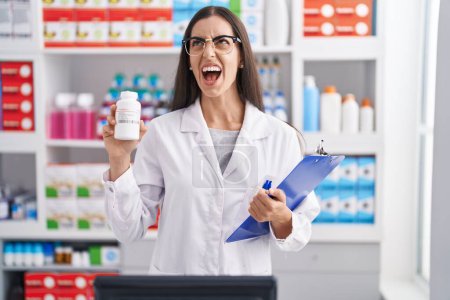 Photo for Young brunette woman working at pharmacy drugstore holding pills angry and mad screaming frustrated and furious, shouting with anger. rage and aggressive concept. - Royalty Free Image
