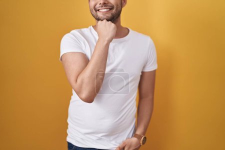 Foto de Hispanic man wearing white t shirt over yellow background serious face thinking about question with hand on chin, thoughtful about confusing idea - Imagen libre de derechos