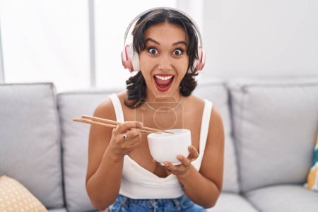 Téléchargez les photos : Young hispanic woman eating asian food using chopsticks celebrating crazy and amazed for success with open eyes screaming excited. - en image libre de droit