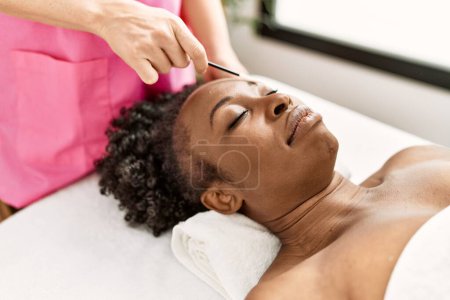 Téléchargez les photos : African american woman lying on massage table having eyebrows treatment at beauty salon - en image libre de droit