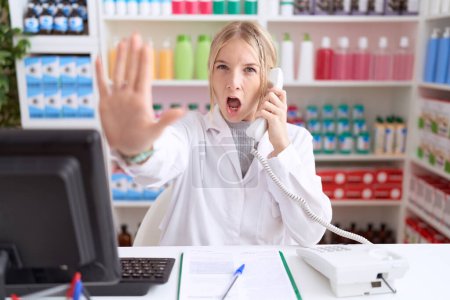 Téléchargez les photos : Jeune femme caucasienne travaillant à la pharmacie pharmacie parlant au téléphone faisant geste d'arrêt avec les paumes des mains, expression de colère et de frustration - en image libre de droit