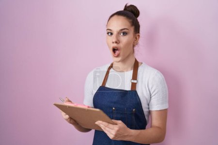 Téléchargez les photos : Young hispanic girl wearing professional waitress apron taking order afraid and shocked with surprise and amazed expression, fear and excited face. - en image libre de droit