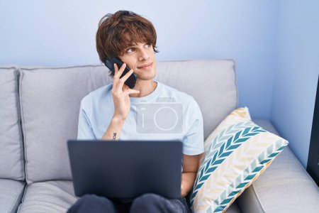 Téléchargez les photos : Young blond man talking on smartphone using laptop at home - en image libre de droit