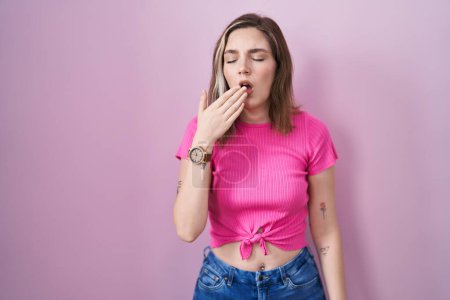 Foto de Blonde caucasian woman standing over pink background bored yawning tired covering mouth with hand. restless and sleepiness. - Imagen libre de derechos