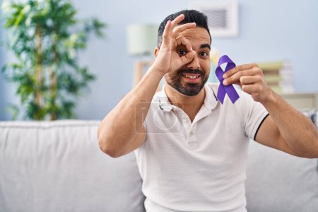 Téléchargez les photos : Young hispanic man with beard holding purple ribbon awareness smiling happy doing ok sign with hand on eye looking through fingers - en image libre de droit