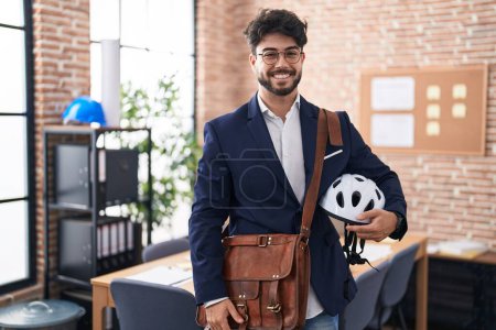 Téléchargez les photos : Hispanic man with beard holding bike helmet at the office looking positive and happy standing and smiling with a confident smile showing teeth - en image libre de droit