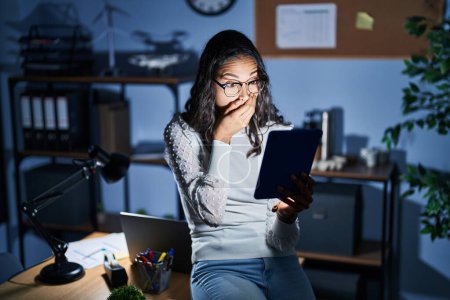 Téléchargez les photos : Young brazilian woman using touchpad at night working at the office shocked covering mouth with hands for mistake. secret concept. - en image libre de droit