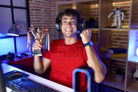 Photo for Young hispanic man playing video games holding trophy screaming proud, celebrating victory and success very excited with raised arm - Royalty Free Image