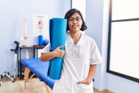 Photo for Young latin woman wearing physiotherapist uniform holding yoga mat at physiotherapy clinic - Royalty Free Image