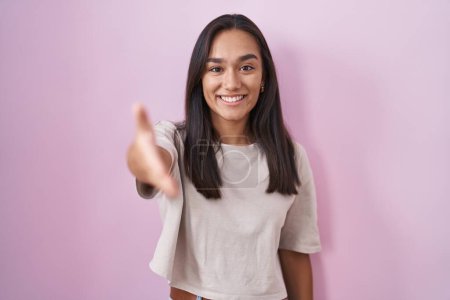 Foto de Young hispanic woman standing over pink background smiling friendly offering handshake as greeting and welcoming. successful business. - Imagen libre de derechos
