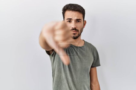 Foto de Young hispanic man with beard wearing casual t shirt over white background looking unhappy and angry showing rejection and negative with thumbs down gesture. bad expression. - Imagen libre de derechos
