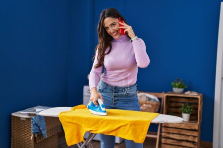 Foto de Young hispanic woman talking on the smartphone ironing clothes at laundry room - Imagen libre de derechos