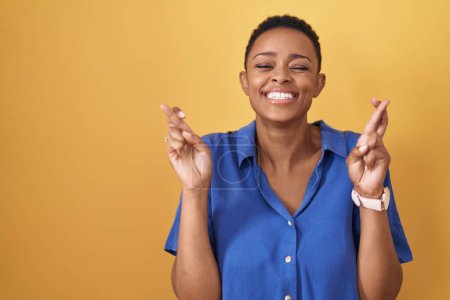 Téléchargez les photos : Femme afro-américaine debout sur fond jaune geste doigt croisé souriant avec espoir et les yeux fermés. chance et concept superstitieux. - en image libre de droit