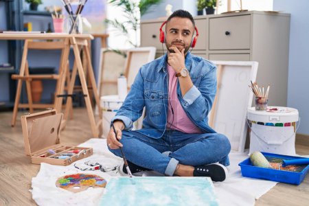 Téléchargez les photos : Jeune peintre hispanique assis sur le sol à l'atelier d'art visage sérieux penser à la question avec la main sur le menton, réfléchi à l'idée déroutante - en image libre de droit
