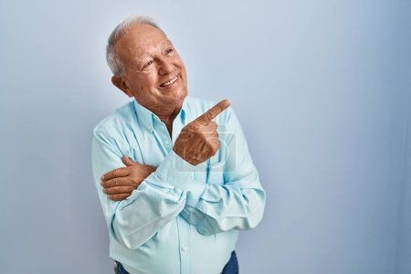 Foto de Hombre mayor con el pelo gris de pie sobre fondo azul con una gran sonrisa en la cara, apuntando con la mano y el dedo hacia el lado mirando a la cámara. - Imagen libre de derechos