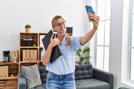 Téléchargez les photos : Jeune femme blonde psychologue souriant confiant faire selfie par le smartphone à la clinique - en image libre de droit