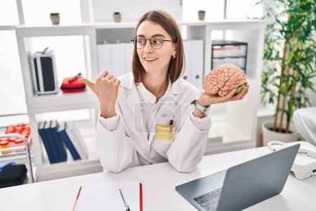 Téléchargez les photos : Jeune médecin caucasien femme tenant le cerveau comme concept de santé mentale pointant le pouce vers le côté souriant heureux avec la bouche ouverte - en image libre de droit
