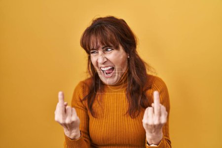 Foto de Middle age hispanic woman standing over yellow background showing middle finger doing fuck you bad expression, provocation and rude attitude. screaming excited - Imagen libre de derechos