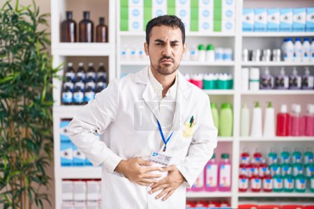 Téléchargez les photos : Handsome hispanic man working at pharmacy drugstore with hand on stomach because indigestion, painful illness feeling unwell. ache concept. - en image libre de droit