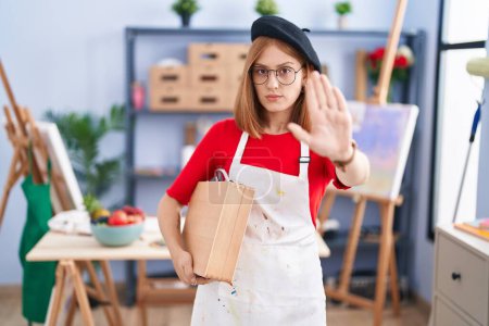 Photo for Young redhead woman at art studio holding art case doing stop sing with palm of the hand. warning expression with negative and serious gesture on the face. - Royalty Free Image