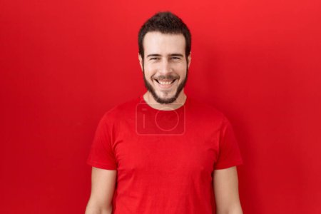 Foto de Joven hombre hispano vistiendo una camiseta roja casual con una sonrisa alegre y fresca en la cara. persona afortunada. - Imagen libre de derechos