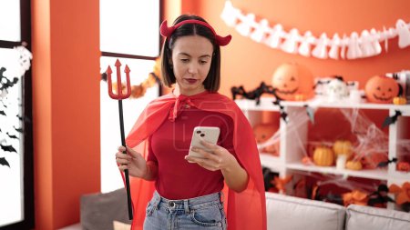 Téléchargez les photos : Young beautiful hispanic woman wearing devil costume using smartphone at home - en image libre de droit