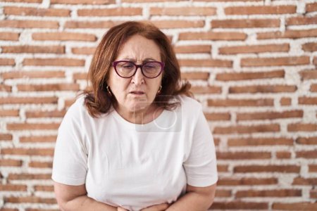 Foto de Senior woman with glasses standing over bricks wall with hand on stomach because indigestion, painful illness feeling unwell. ache concept. - Imagen libre de derechos